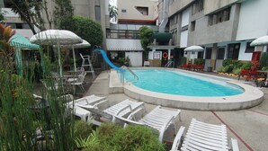 A swimming pool with a slide, surrounded by lounge chairs and umbrellas, set in a courtyard with buildings in the background. - Hotel Supreme (Baguio)