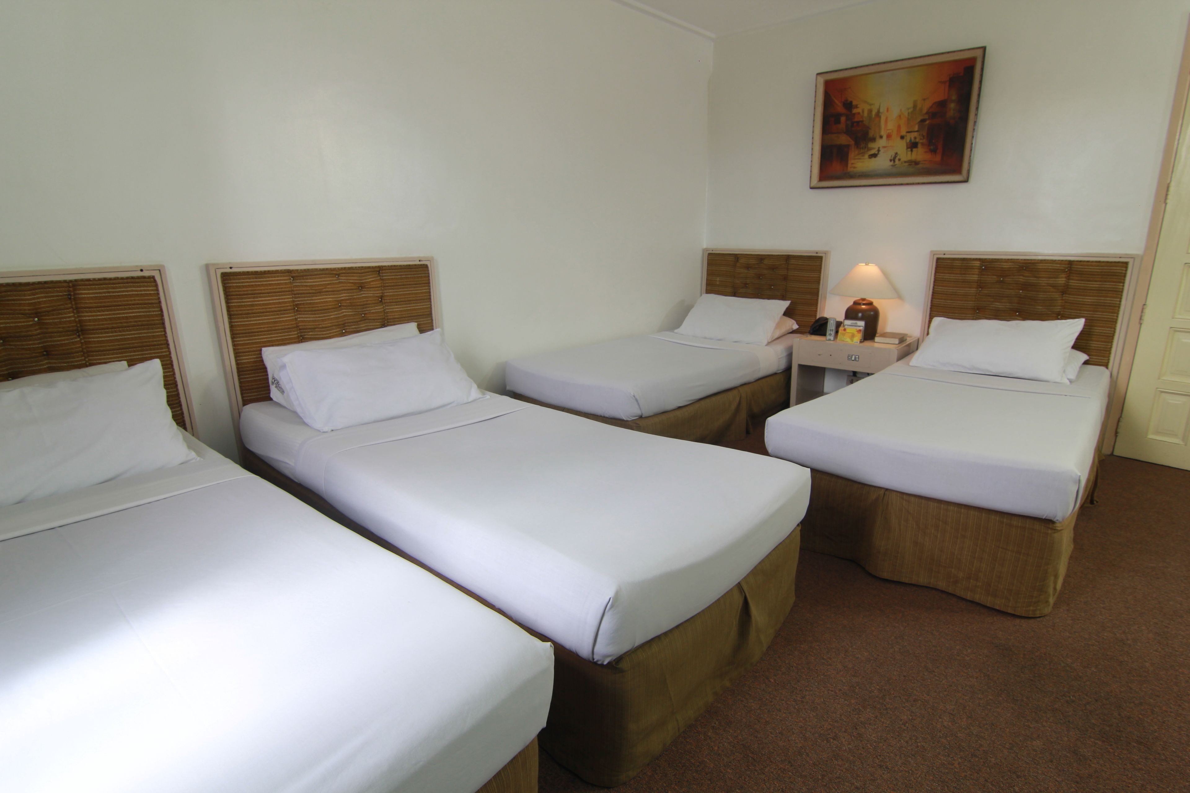 Une chambre d’hôtel avec trois lits simples, chacun doté d’une tête de lit en bois et de draps blancs. Il y a une table de chevet avec une lampe et une petite table entre les lits. Un tableau est accroché au mur, au-dessus des lits.
