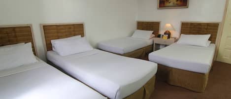 Habitación de hotel con tres camas individuales, cada una con cabecera de madera y ropa de cama blanca. Hay una mesita de noche con una lámpara y una pequeña mesa entre las camas. Un cuadro cuelga en la pared sobre las camas.