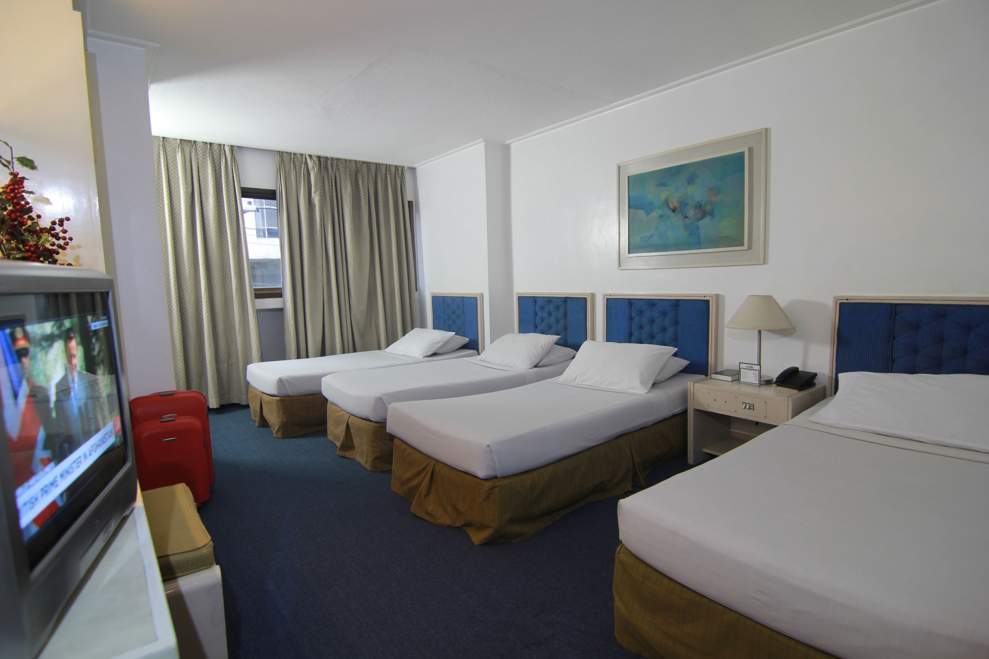 Habitación de hotel con tres camas individuales, televisión, escritorio con lámpara y un cuadro en la pared.