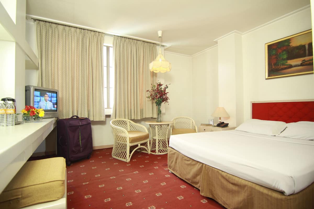 A hotel room with a bed, a desk with a television, a chair, a small table with a vase, and a painting on the wall.