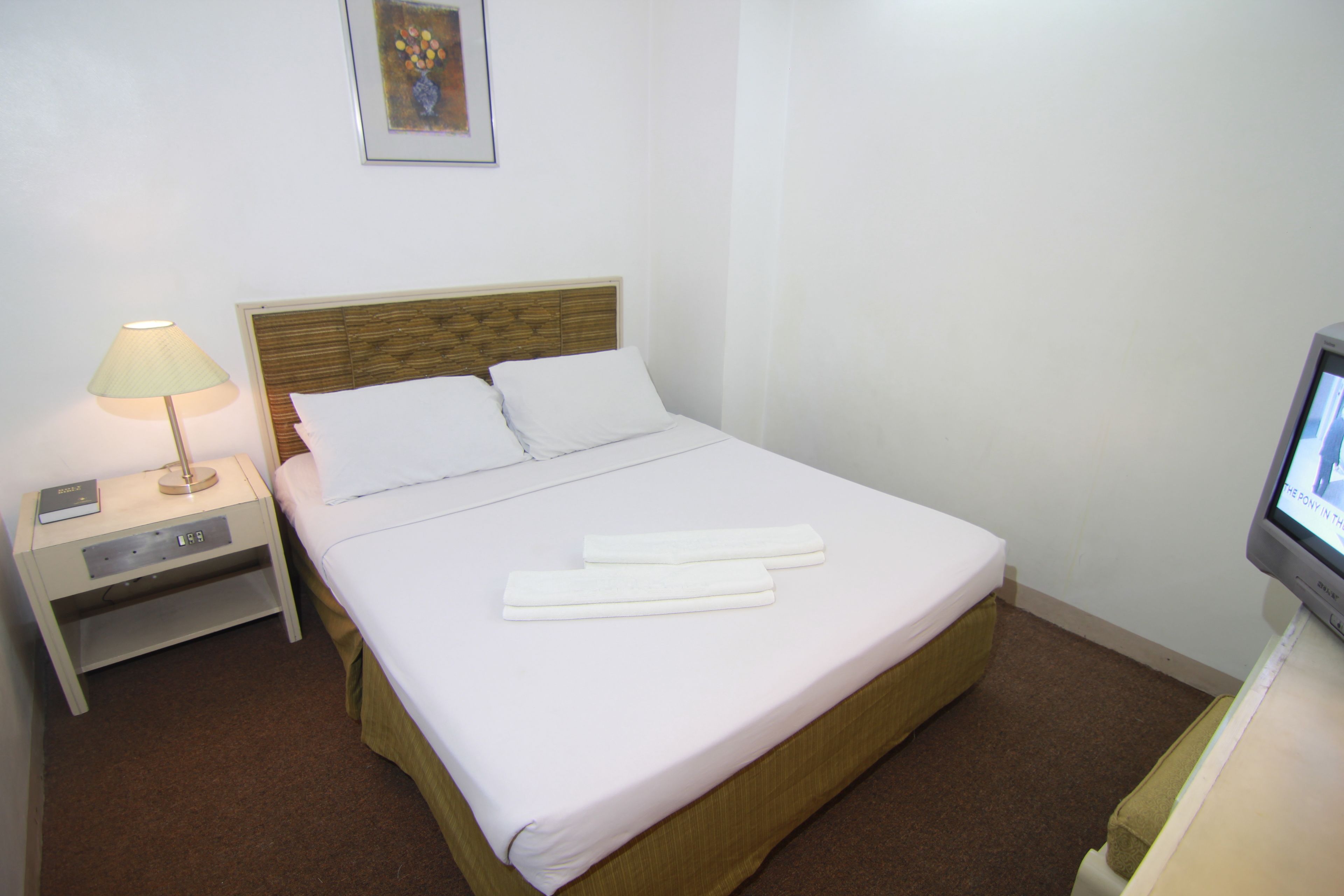 Una cama bien hecha con ropa de cama y toallas blancas, una lámpara de noche, un estante pequeño y un televisor sobre un mueble bajo.