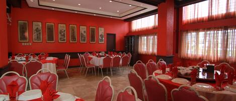 Un salón de banquetes con paredes rojas, varias mesas redondas dispuestas para comer, y cuadros enmarcados en las paredes.