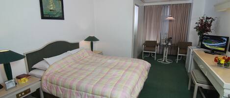 A hotel room with a bed, a desk with a computer and a chair, a TV, and a painting on the wall.