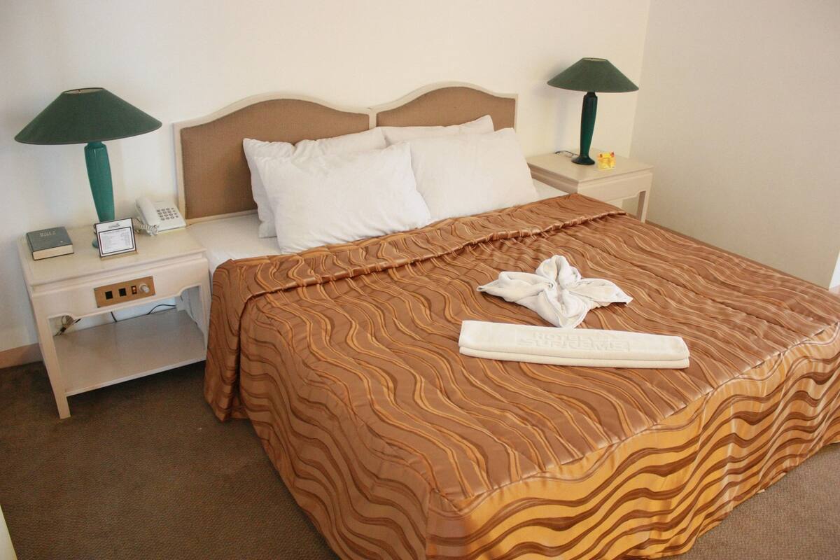 A neatly made bed with a brown patterned bedspread, two white pillows, and a folded towel on the bed. There are two bedside tables with lamps and a phone on one of them.