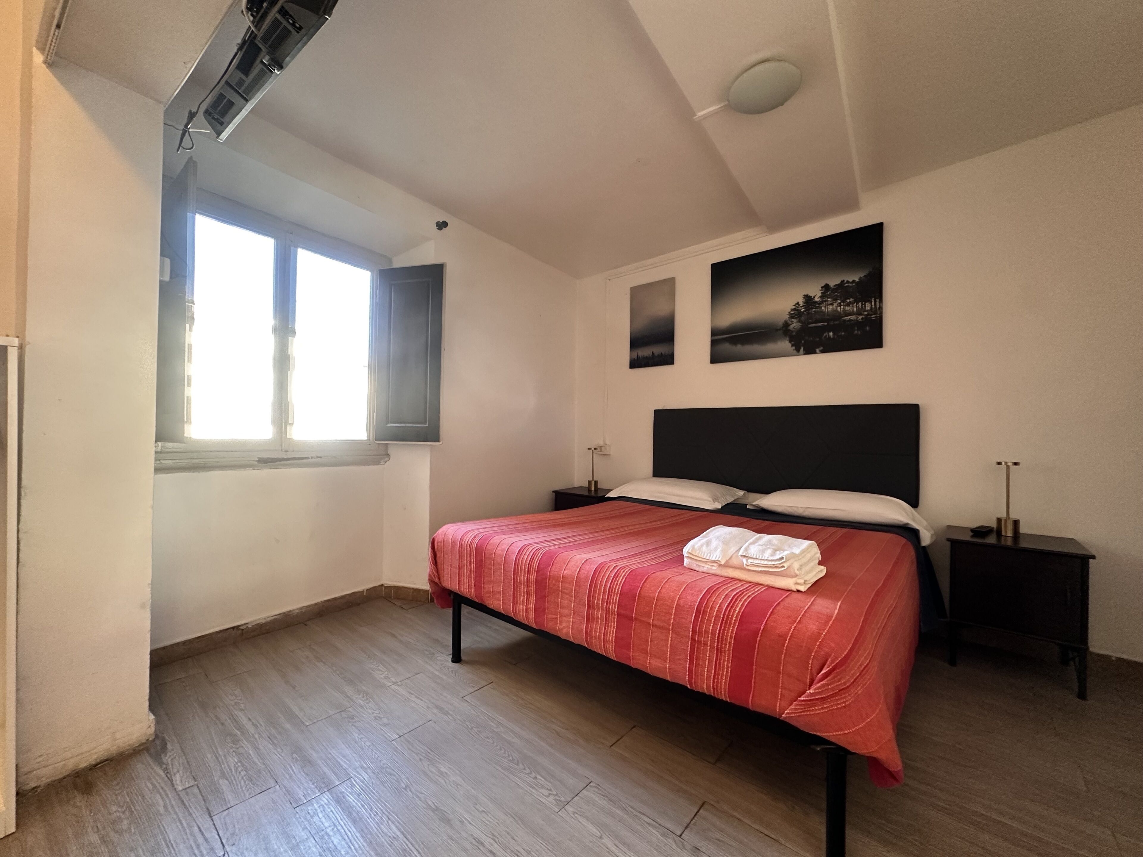 A bedroom with a bed, two bedside tables, a window, and two wall-mounted pictures.
