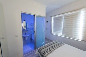 Un dormitorio con cama, un baño con lavamanos e inodoro, y una ventana con persianas.