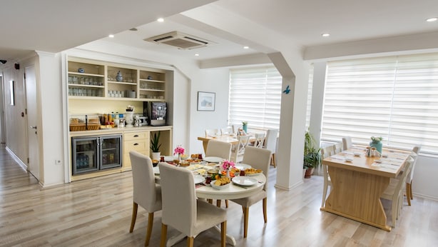 Una cocina moderna con estantes integrados, un comedor con mesa y sillas de madera, y un amplio ventanal con persianas.