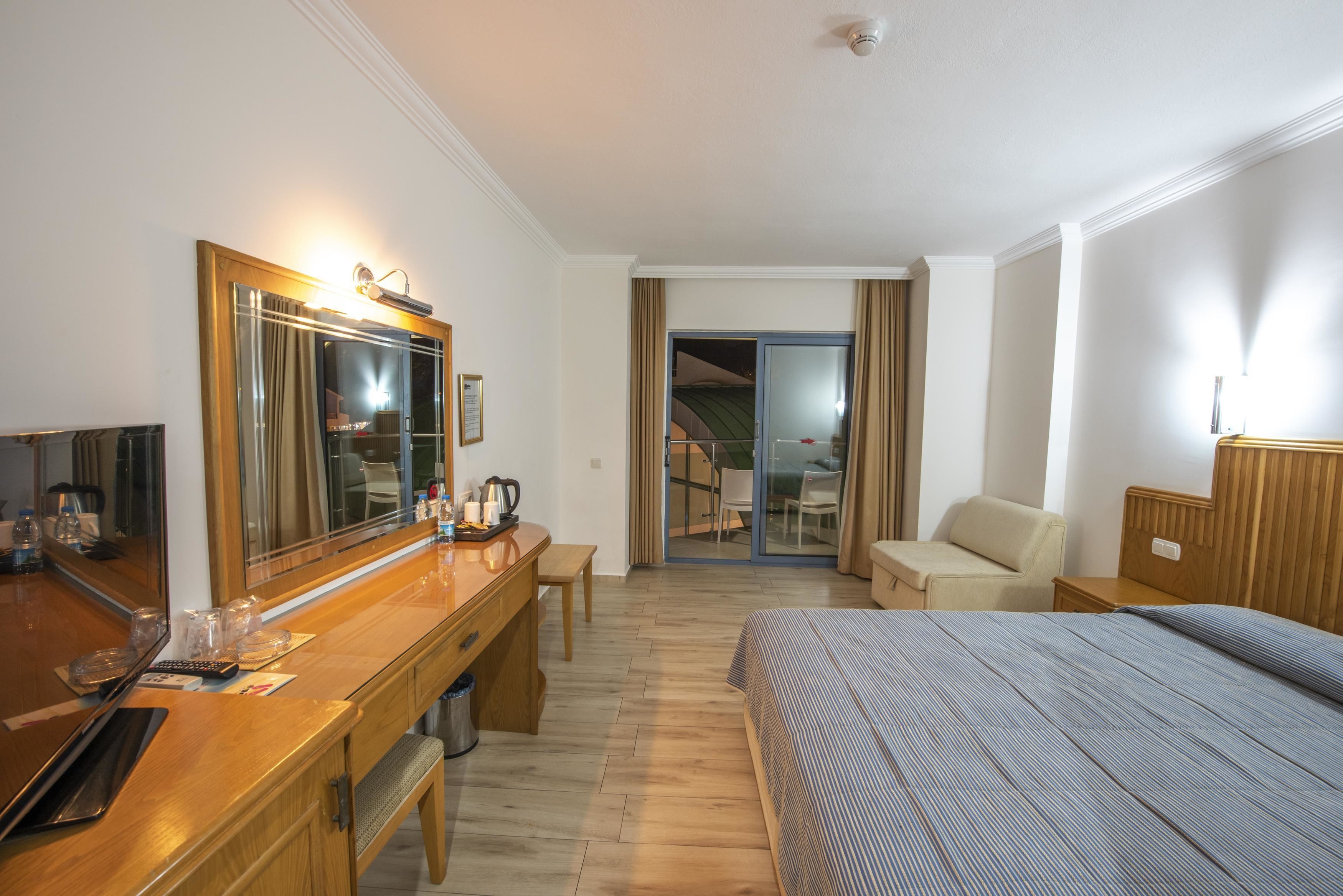 A hotel room with a wooden desk, a bed with a striped bedspread, a large mirror, and a sliding glass door leading to a balcony.