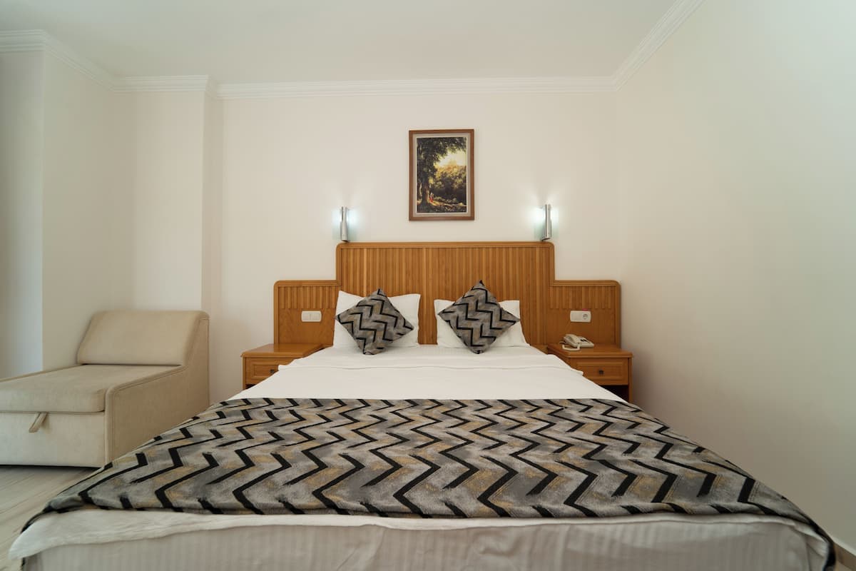 A hotel room with a bed, a nightstand, a lamp, a chair, and a painting on the wall.
