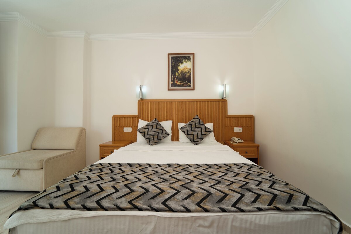 A hotel room with a bed, a nightstand, a lamp, a chair, and a painting on the wall.