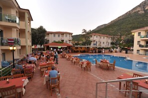 Zona de piscina al aire libre con varias mesas y sillas para comer, rodeada de edificios con balcones y un paisaje montañoso de fondo.