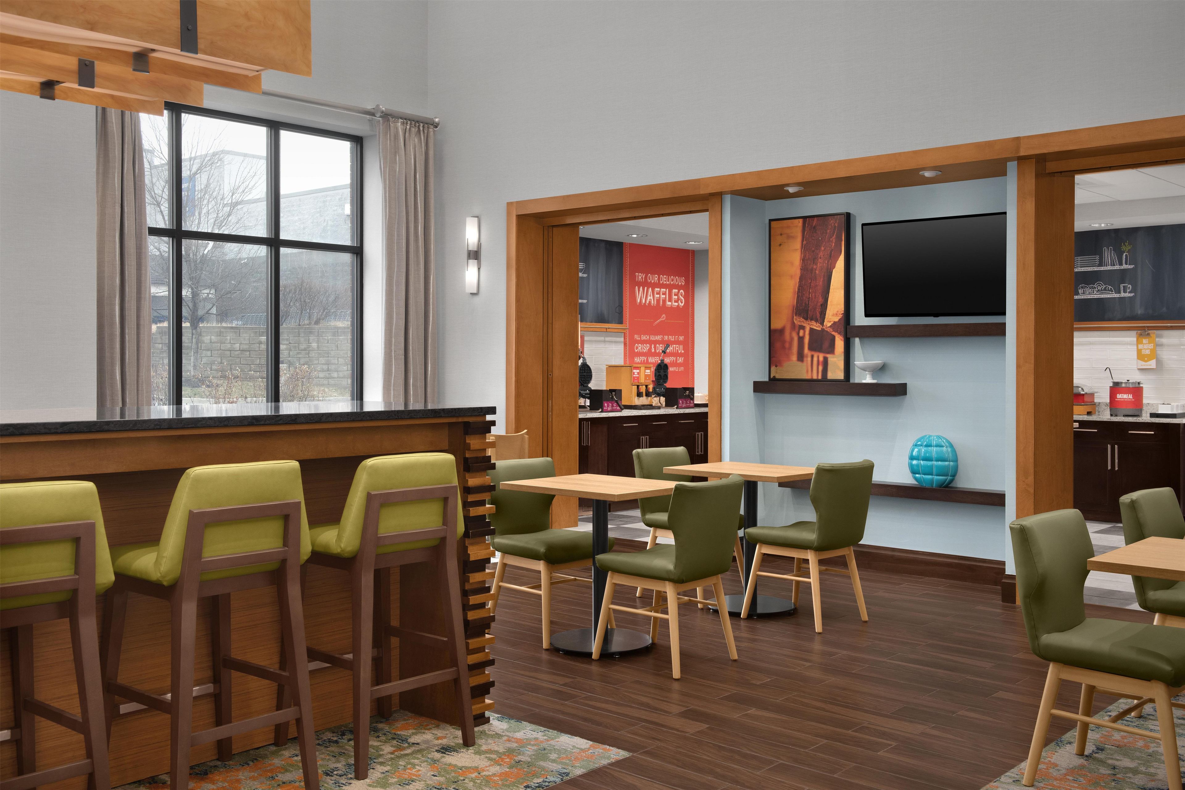 Un área de comedor moderna con mesas y sillas de madera, una cocina al fondo y un ventanal grande.