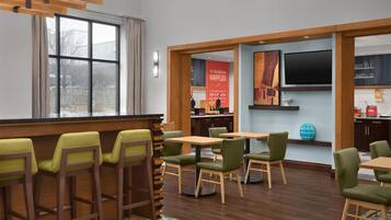 Un área de comedor moderna con mesas y sillas de madera, una cocina al fondo y un ventanal grande.