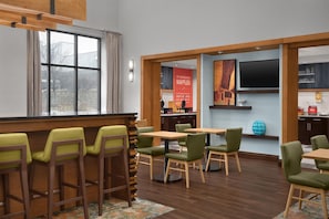 A modern dining area with wooden tables and chairs, a kitchen in the background, and a large window.