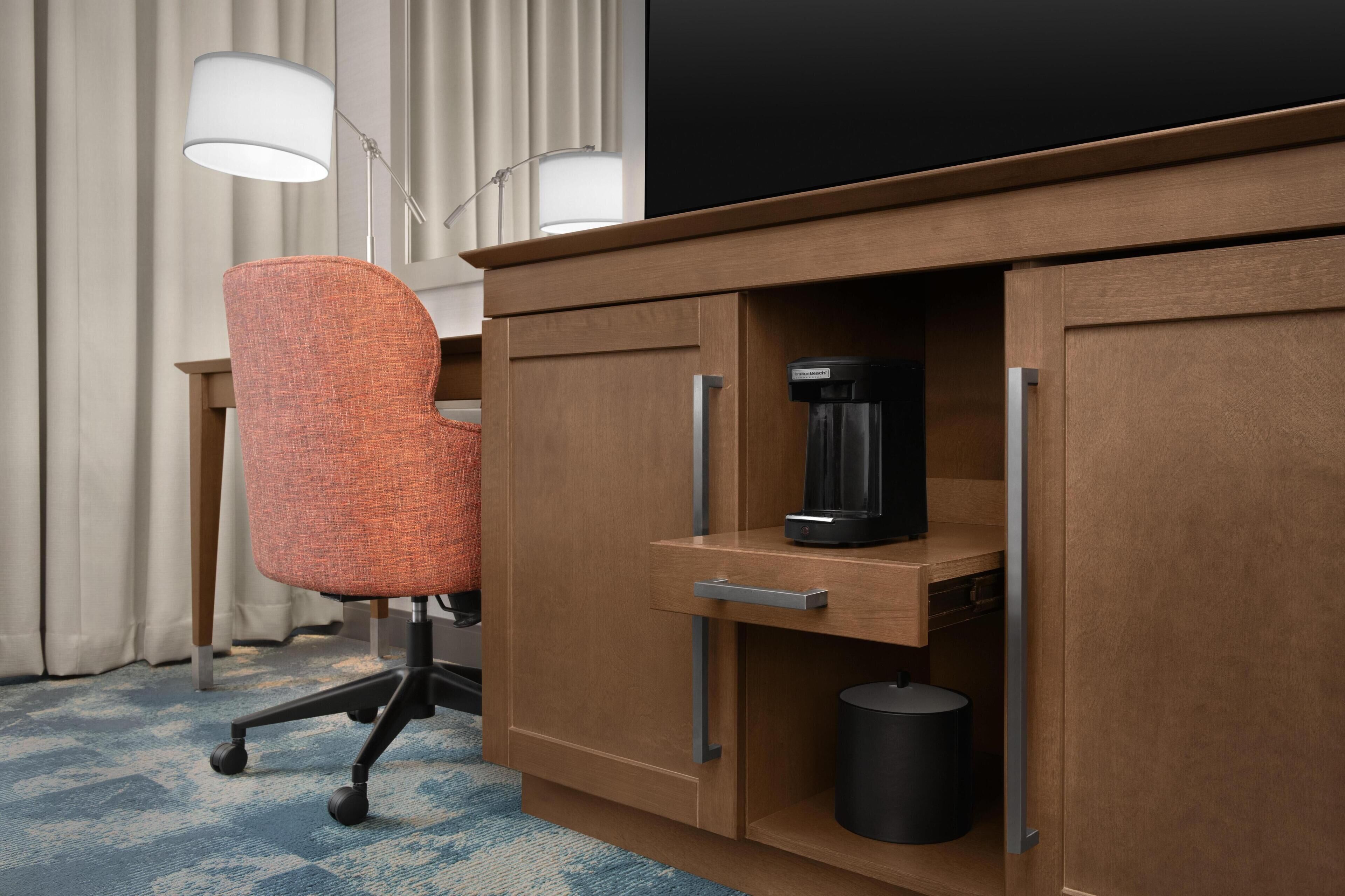 Habitación de hotel con escritorio de madera, una silla de oficina naranja, cafetera y cubo de basura.