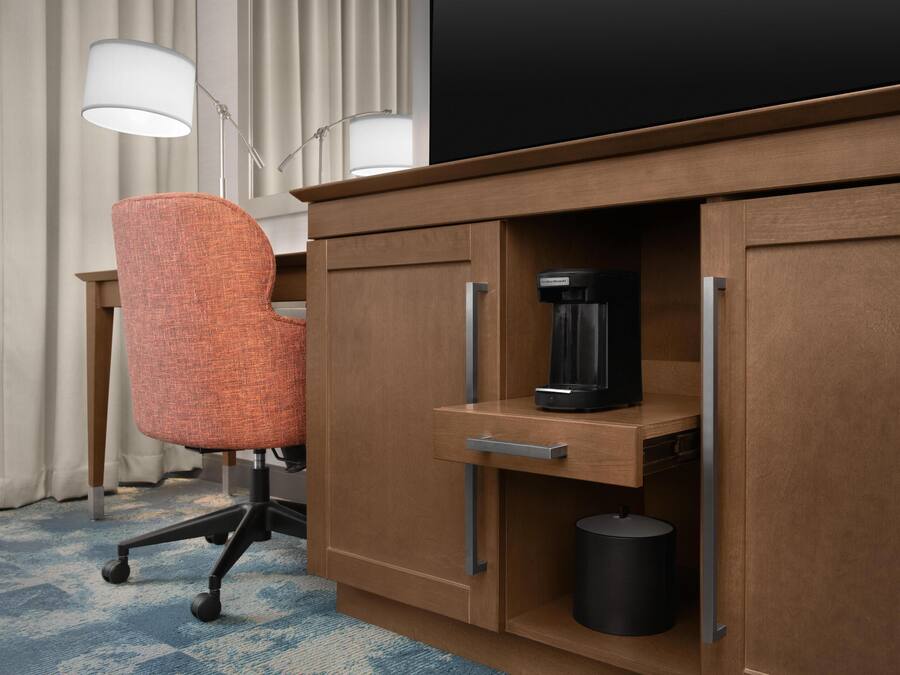 A hotel room with a wooden desk, an orange office chair, a coffee maker, and a trash can.