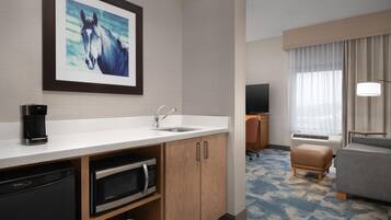 Habitación de hotel con kitchenette, televisor montado, sofá y ventana con cortinas.
