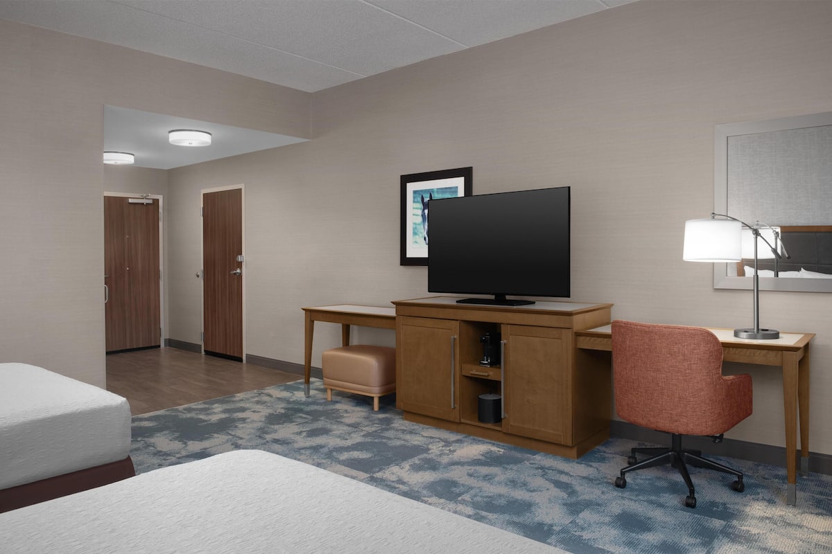 A hotel room with a bed, a desk with a television, a chair, a lamp, and a framed picture on the wall.