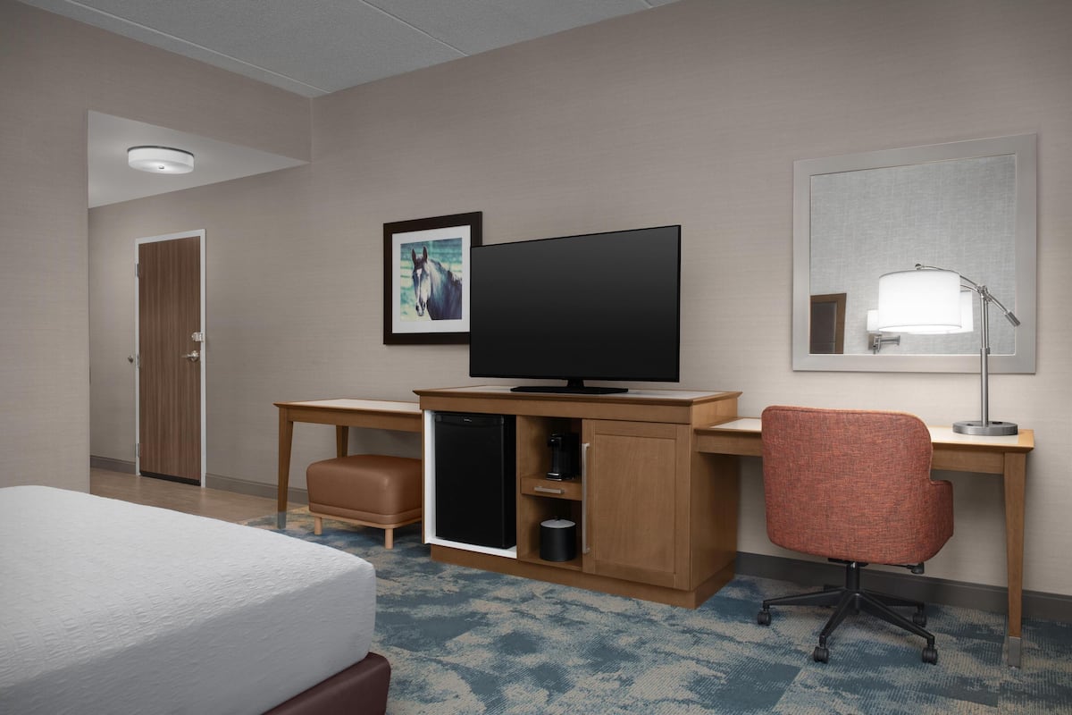 A hotel room with a bed, a desk with a television, a chair, a lamp, and a door.