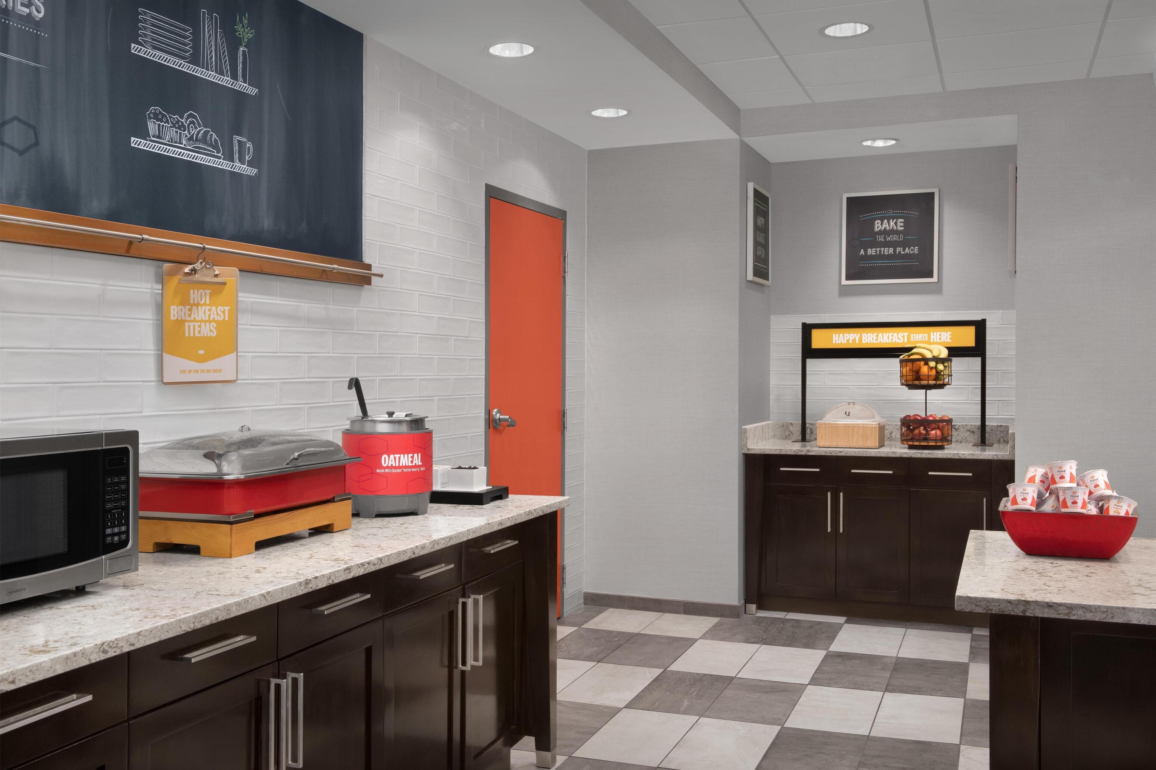 A modern kitchen with a microwave, toaster, and coffee maker on the counter, a red door, and a sign indicating breakfast items.