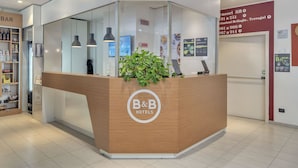 Ein Empfangstresen mit B&B Logo, einer Pflanze und einem Schild mit Wegweisern zu verschiedenen Hotelbereichen.