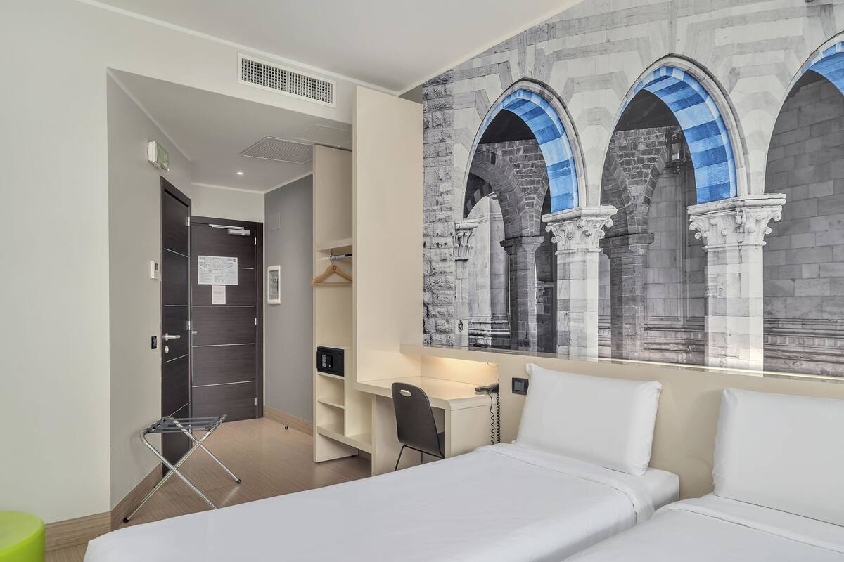 A hotel room with a bed, a desk, a chair, a wardrobe, and a wall with a stone arch design.