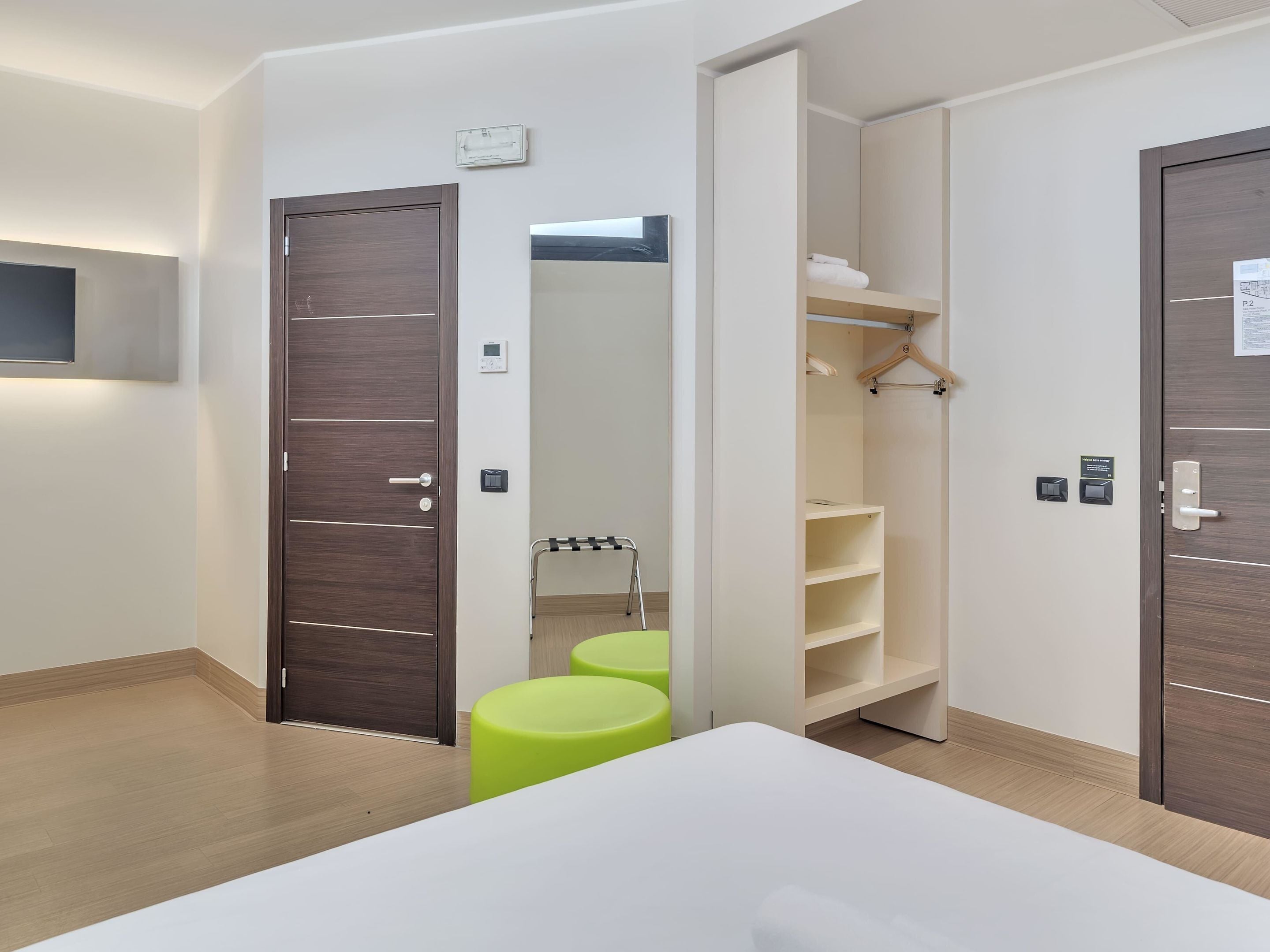 Una habitaci ón de hotel moderna con una cama grande, un armario con espejo, un pequeño rincón de descanso y un televisor integrado.
