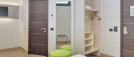 A modern hotel room with a large bed, a wardrobe with a mirror, a small seating area, and a built-in TV.