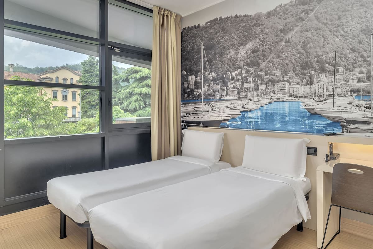 A hotel room with a bed, a desk, and a large window with a view of a marina and a hillside town.