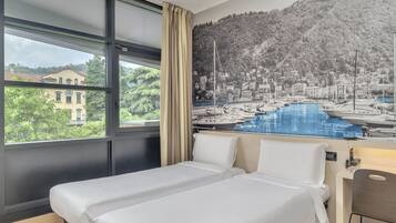 A hotel room with a bed, a desk, and a large window with a view of a marina and a hillside town.