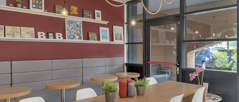 A modern cafe with a long wooden table, white chairs, and a red wall with framed pictures.