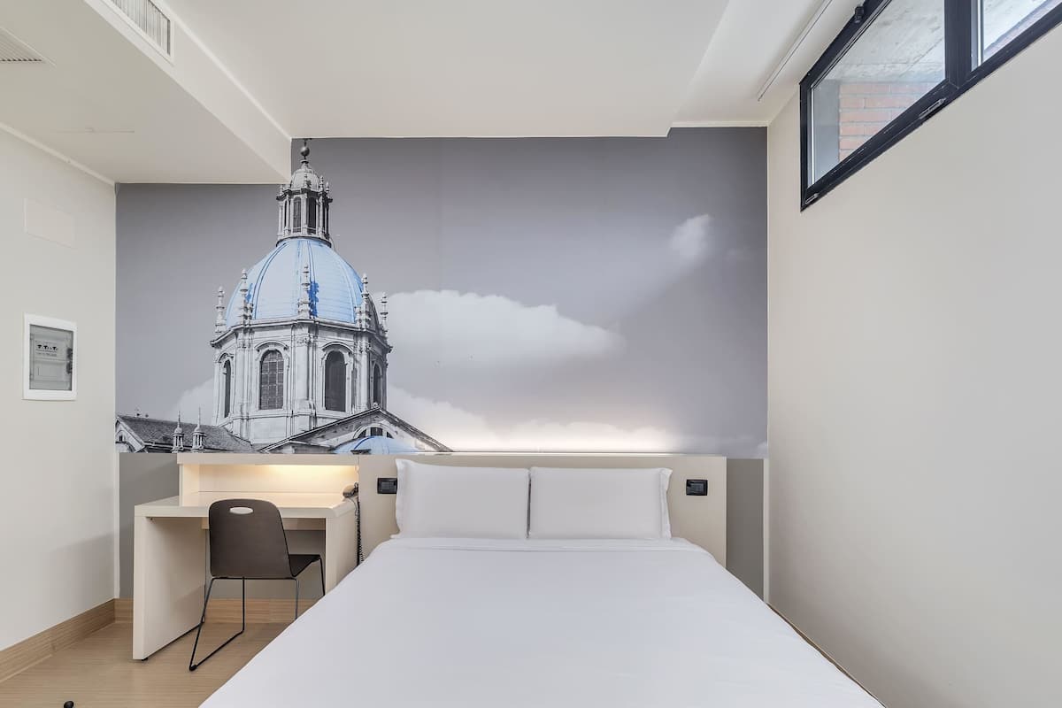 A hotel room with a bed, a desk, and a chair. There is a large wall mural of a building.
