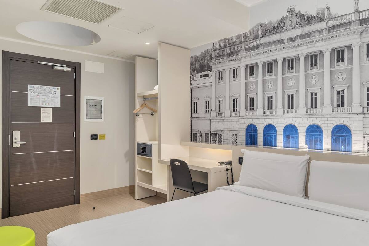 A hotel room with a bed, desk, chair, and a large window with a view of a historic building.