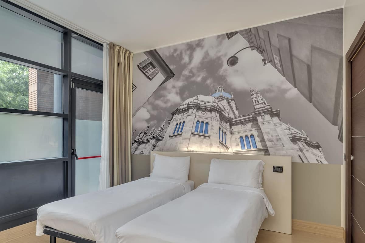 A hotel room with two single beds, a large window with curtains, and a mural of a building on the wall.