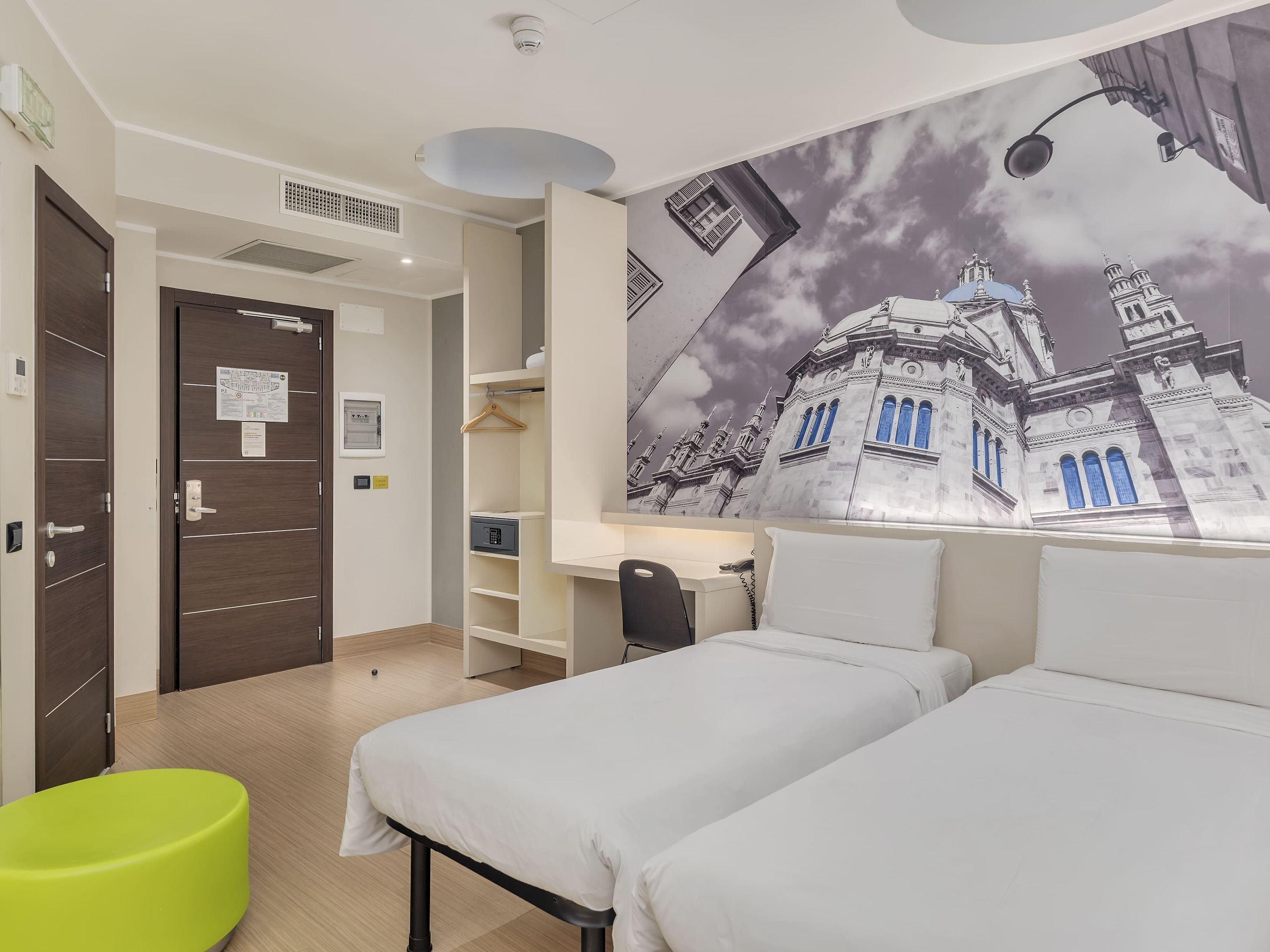 A hotel room with two beds, a desk, a green chair, and a mural of a building.