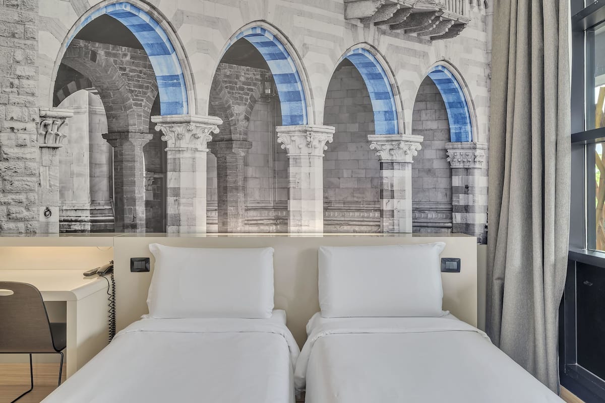 A hotel room with two beds, a desk, and a large window.