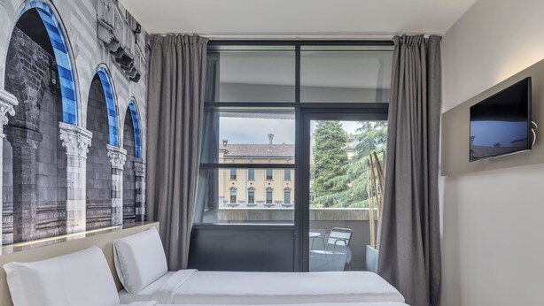 Ein Hotelzimmer mit einem großen Bett, einem an der Wand montierten Fernseher und Blick auf ein Gebäude mit blauen Bögen durch das Fenster.