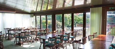 A spacious indoor dining area with wooden tables and chairs, large windows, and a view of greenery outside.