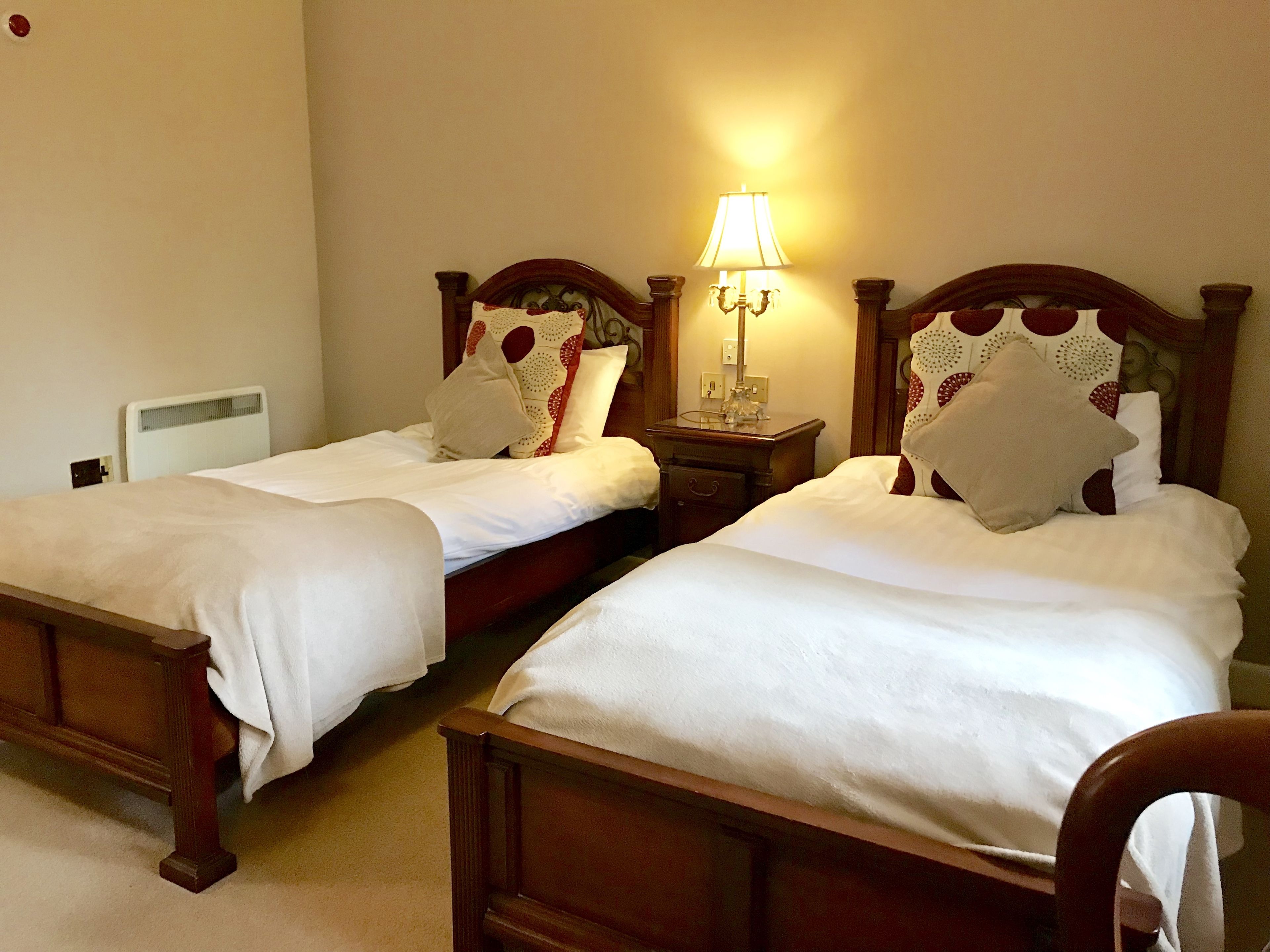 Two single beds with wooden headboards, a bedside table with a lamp, and a radiator on the wall.