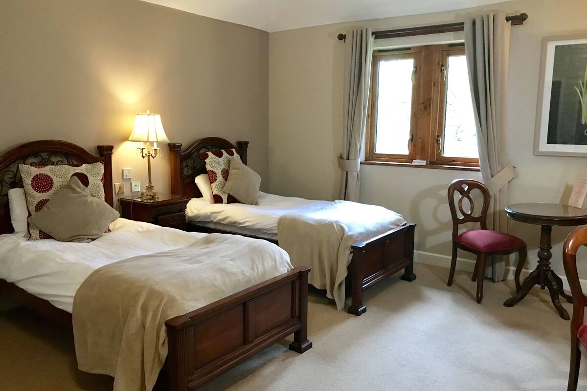 A hotel room with two single beds, a wooden nightstand with a lamp, a wooden chair, a small round table, and a window with curtains.