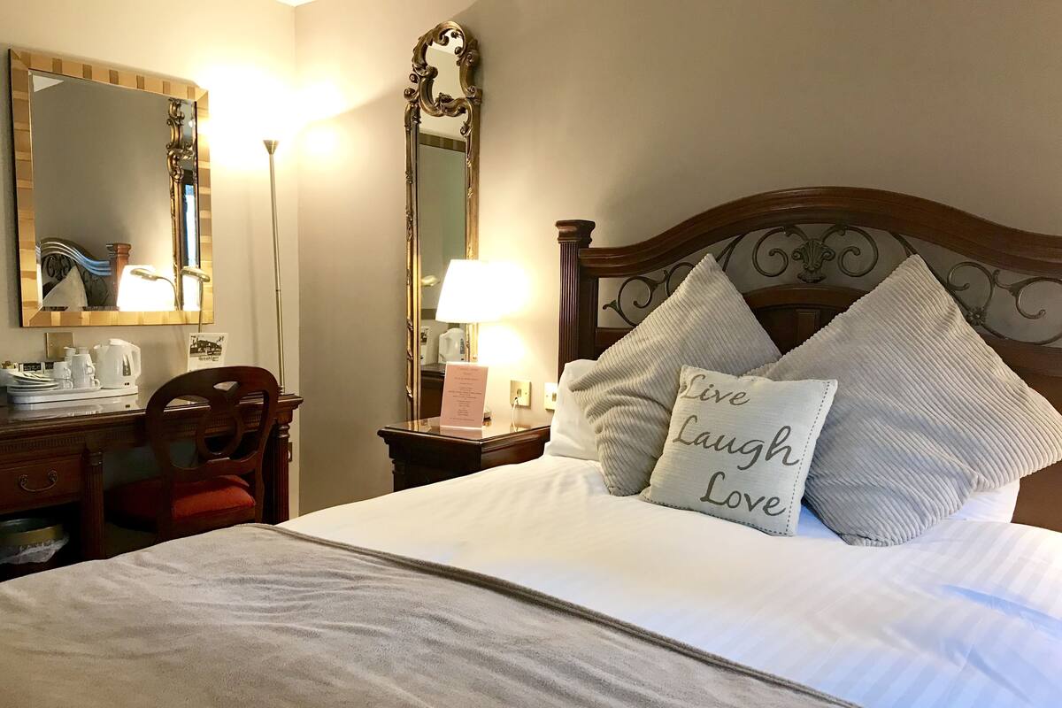 A hotel room with a neatly made bed, a wooden headboard, a desk with a chair, a lamp, and a mirror.