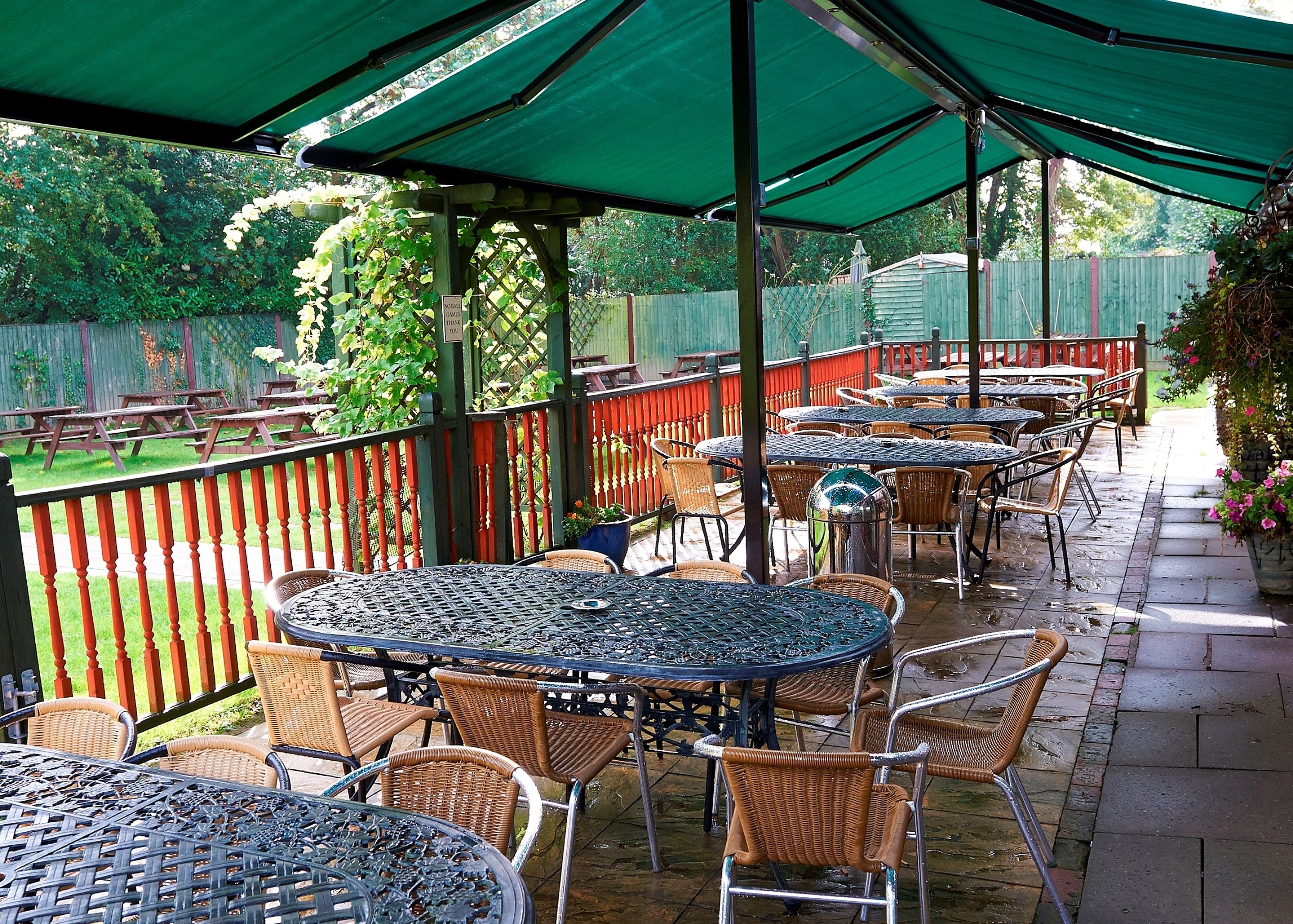 An outdoor patio area with round tables and wicker chairs, covered by green umbrellas, and surrounded by a wooden fence.