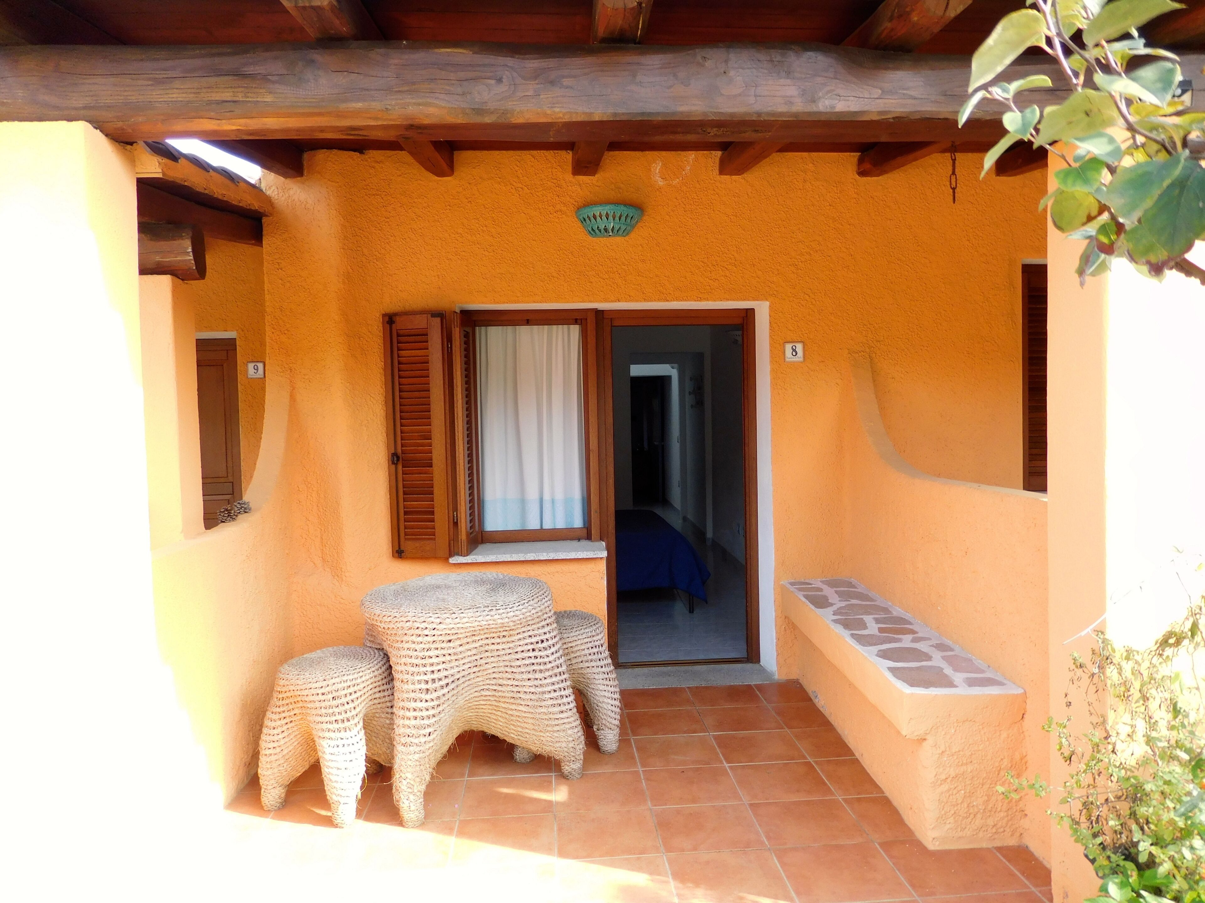 A cozy outdoor seating area with a wooden bench and woven chairs, a door leading inside, and a tiled floor.
