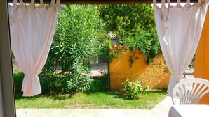 A covered patio with white curtains, a white chair, and a view of a garden with various plants and a yellow wall. - Residence La Pineta (Budoni)