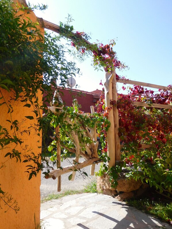 Une pergola en bois ornée de plantes grimpantes et de fleurs, un chemin de pierre et, au fond, un bâtiment avec un toit rouge.