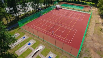 An outdoor tennis court with a green fence, a covered seating area, and a parking lot.