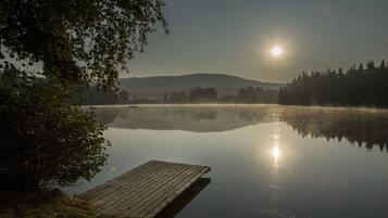 A calm lake with a wooden dock extending into the water, surrounded by trees and a hilly landscape in the background.