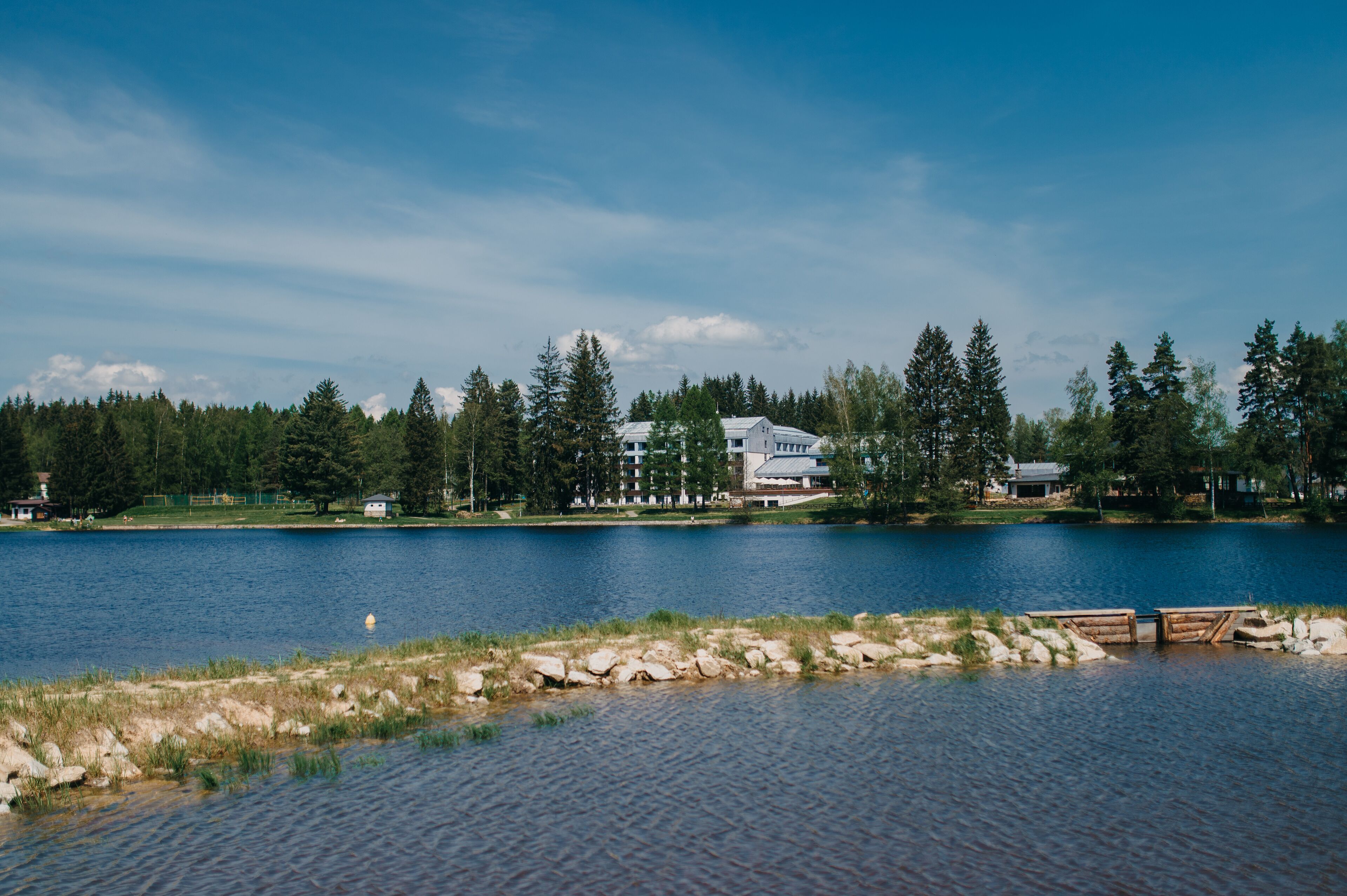 Une vue sur un lac, avec une jetée en bois, un grand bâtiment doté de nombreuses fenêtres et, au loin, une forêt dense.