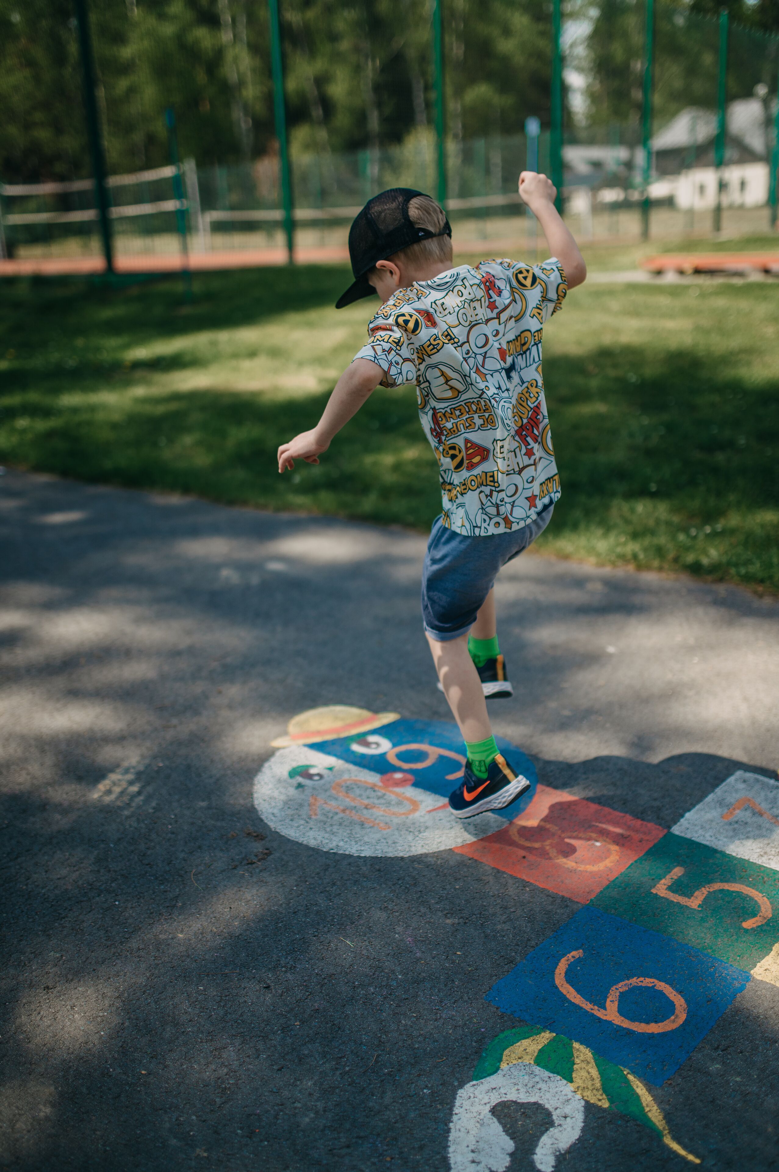 Un bambino gioca a campana sulla scuola in un parco giochi con una recinzione verde e alberi sullo sfondo.